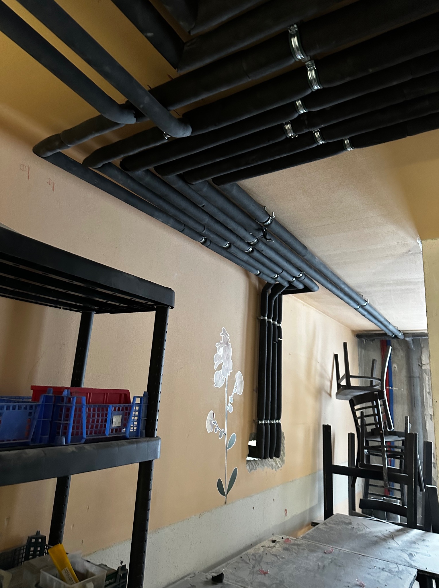 Szereg czarnych, zaizolowanych rur biegnących pod sufitem pomieszczenia, przymocowanych metalowymi obejmami. Na ścianie naklejka w kształcie białego kwiatu. W tle sterta krzeseł i regał z pojemnikami.