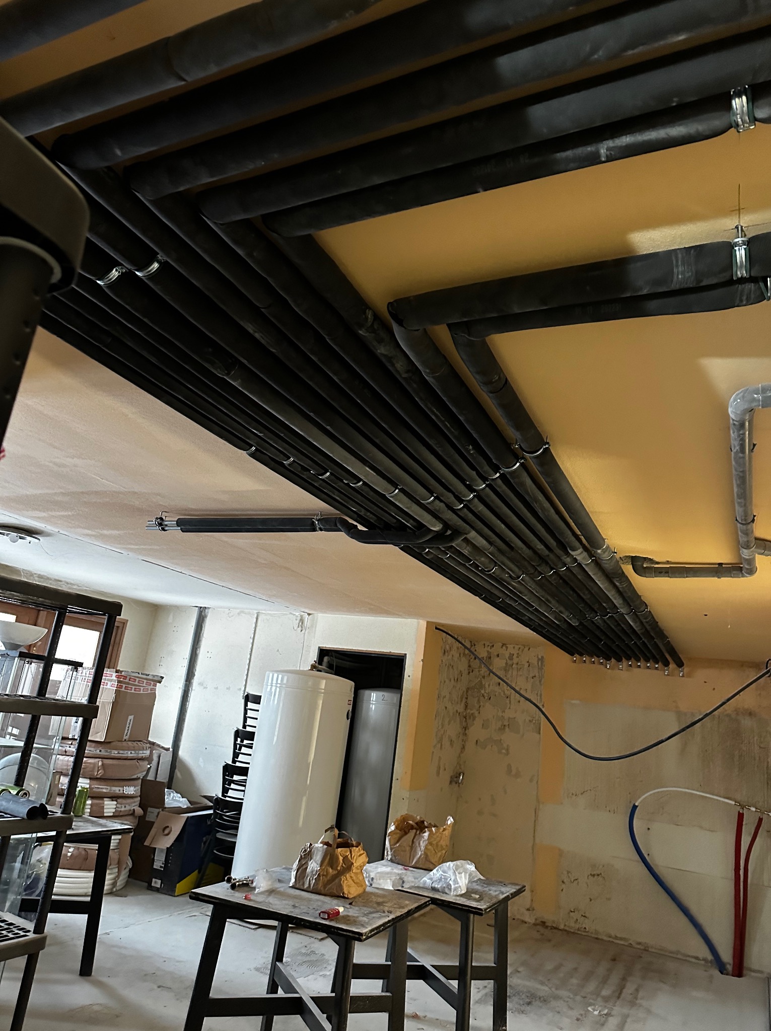 Równoległy układ czarnych rur instalacyjnych pod sufitem w surowym wnętrzu, widoczne uchwyty mocujące, biały bojler i prowizoryczne stanowisko pracy na dwóch stołach.