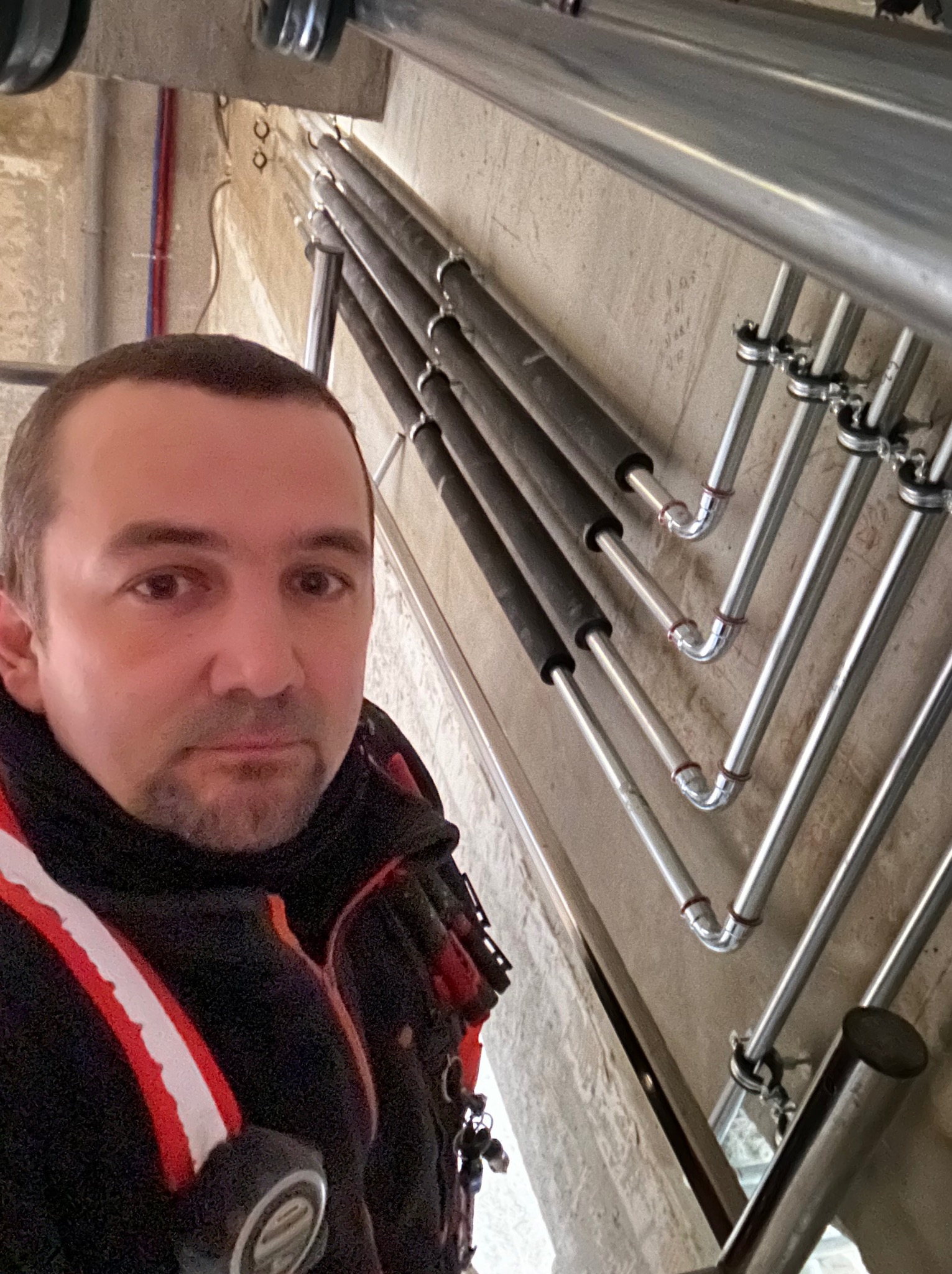 Instalator pozuje na tle pionowo ułożonych rur grzewczych, część z nich zaizolowana czarną pianką, mocowanych do ściany za pomocą metalowych uchwytów.