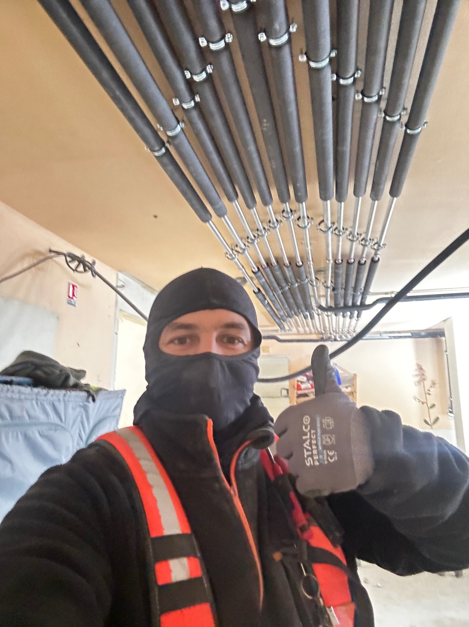 Instalator w kominiarce i odblaskowej kamizelce, z kciukiem w górę, prezentuje rury grzewcze zamontowane pod sufitem w Warszawie. Rury są zabezpieczone obejmami.