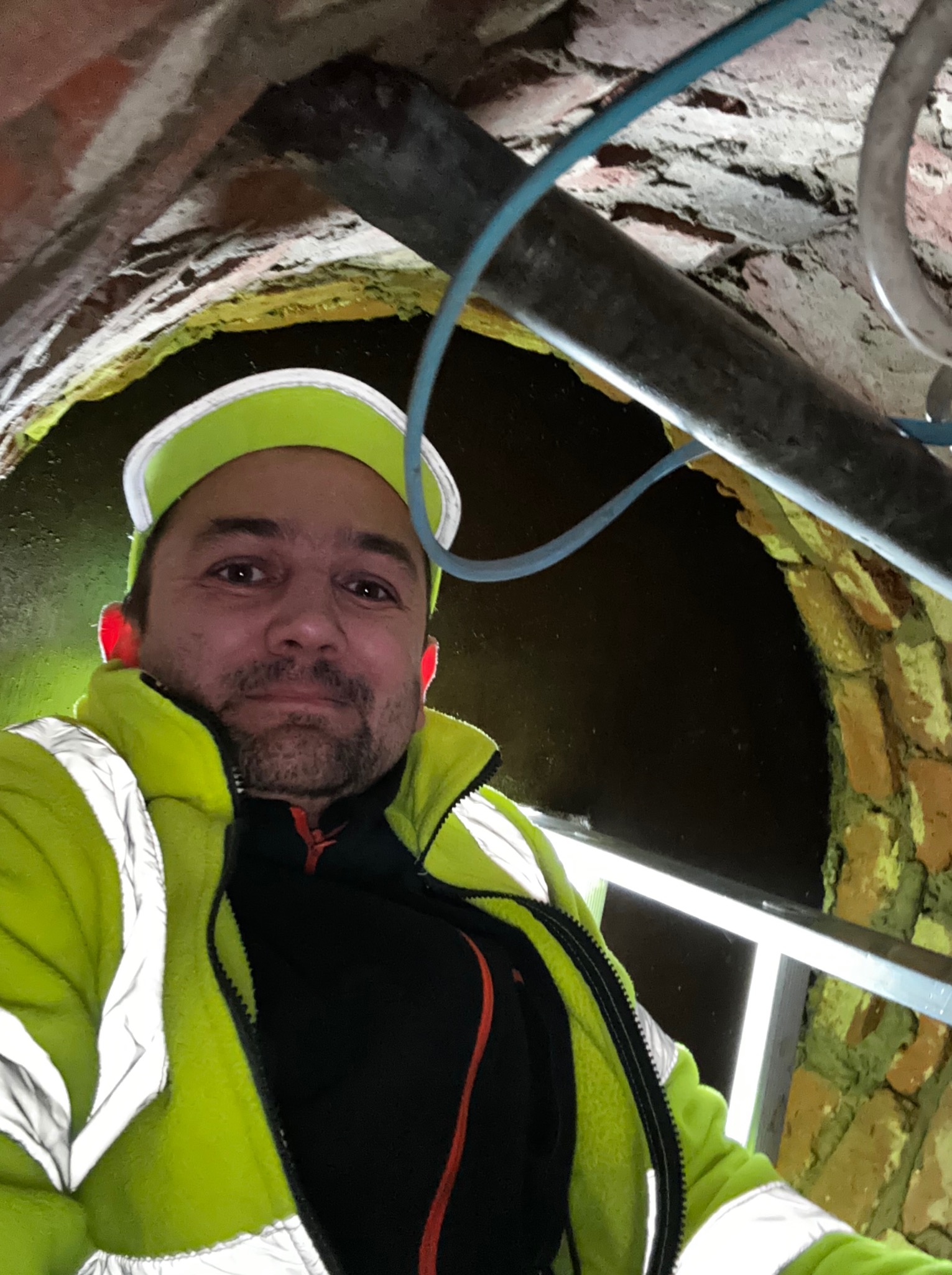 Instalator w odblaskowej kurtce i czapce, robiący selfie wewnątrz ceglanego kanału z rurami i oświetleniem, widok z dołu.
