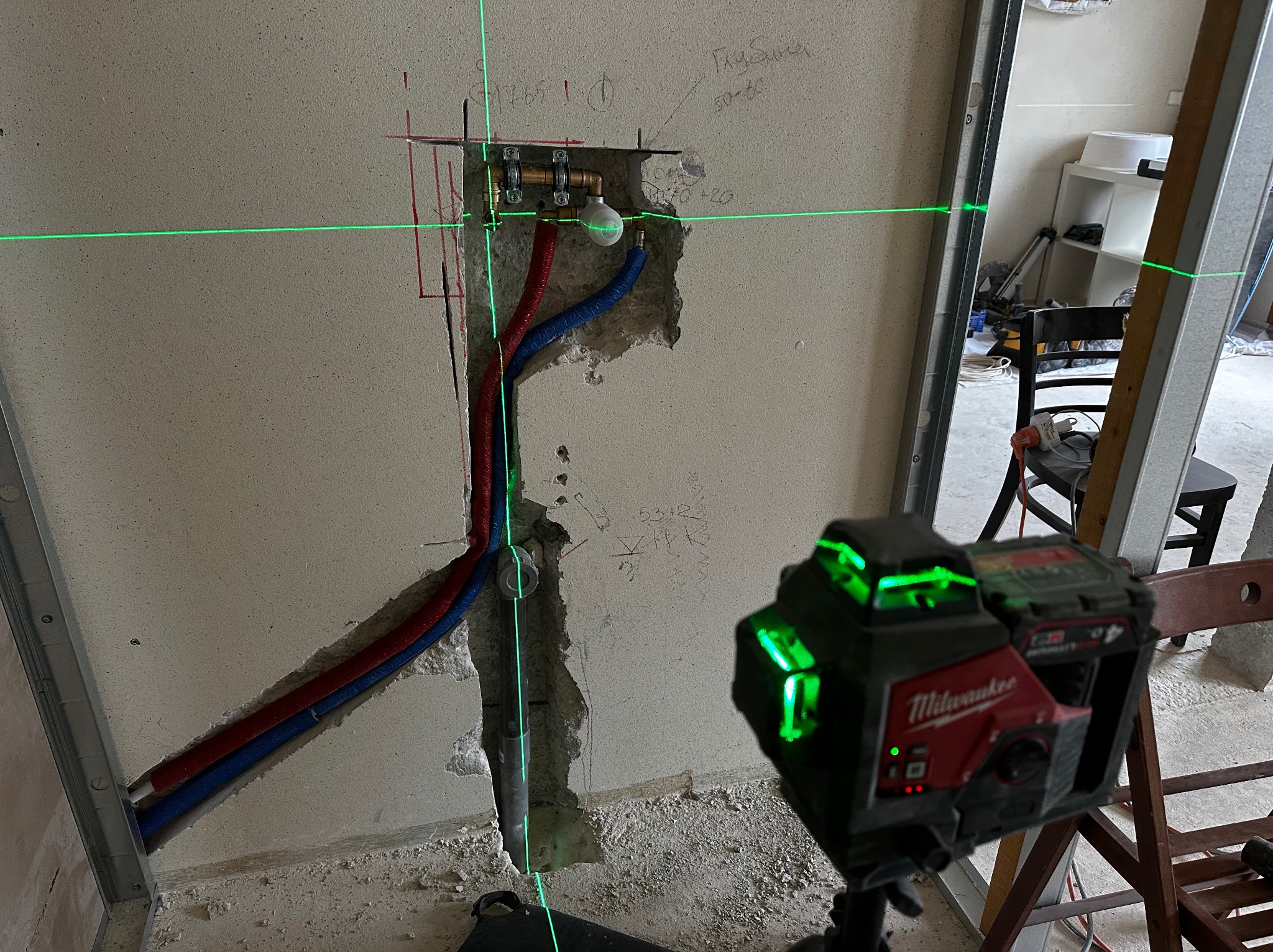 Ściana w trakcie prac instalacyjnych z widocznymi rurami (czerwona i niebieska) oraz elementami mocującymi, zielony laser krzyżowy wyznacza poziom, widoczne notatki na ścianie.