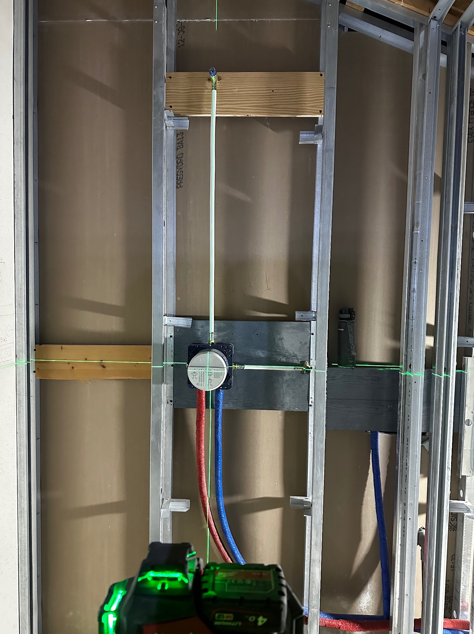 Instalacja podtynkowa baterii Grohe w metalowej konstrukcji szkieletowej, z widocznymi rurami doprowadzającymi wodę (czerwona i niebieska) oraz poziomnicą laserową ustawiającą pion, na tle płyty...