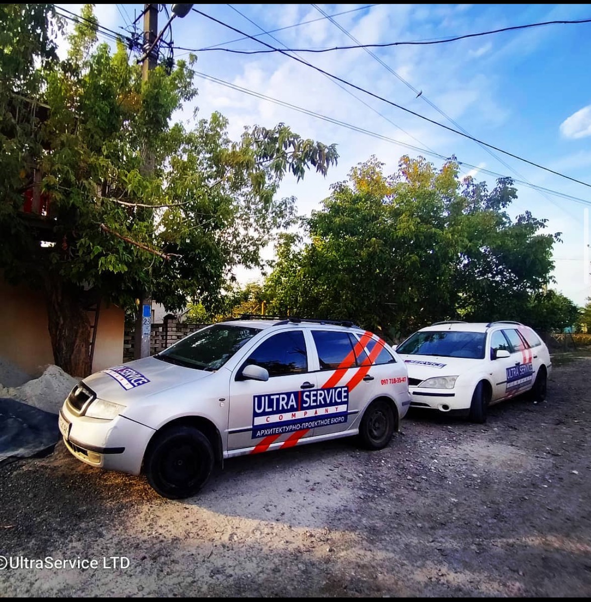 Dwa samochody firmowe z logo 'Ultra Service' zaparkowane na żwirowej drodze, otoczone zielenią, w tle błękitne niebo.