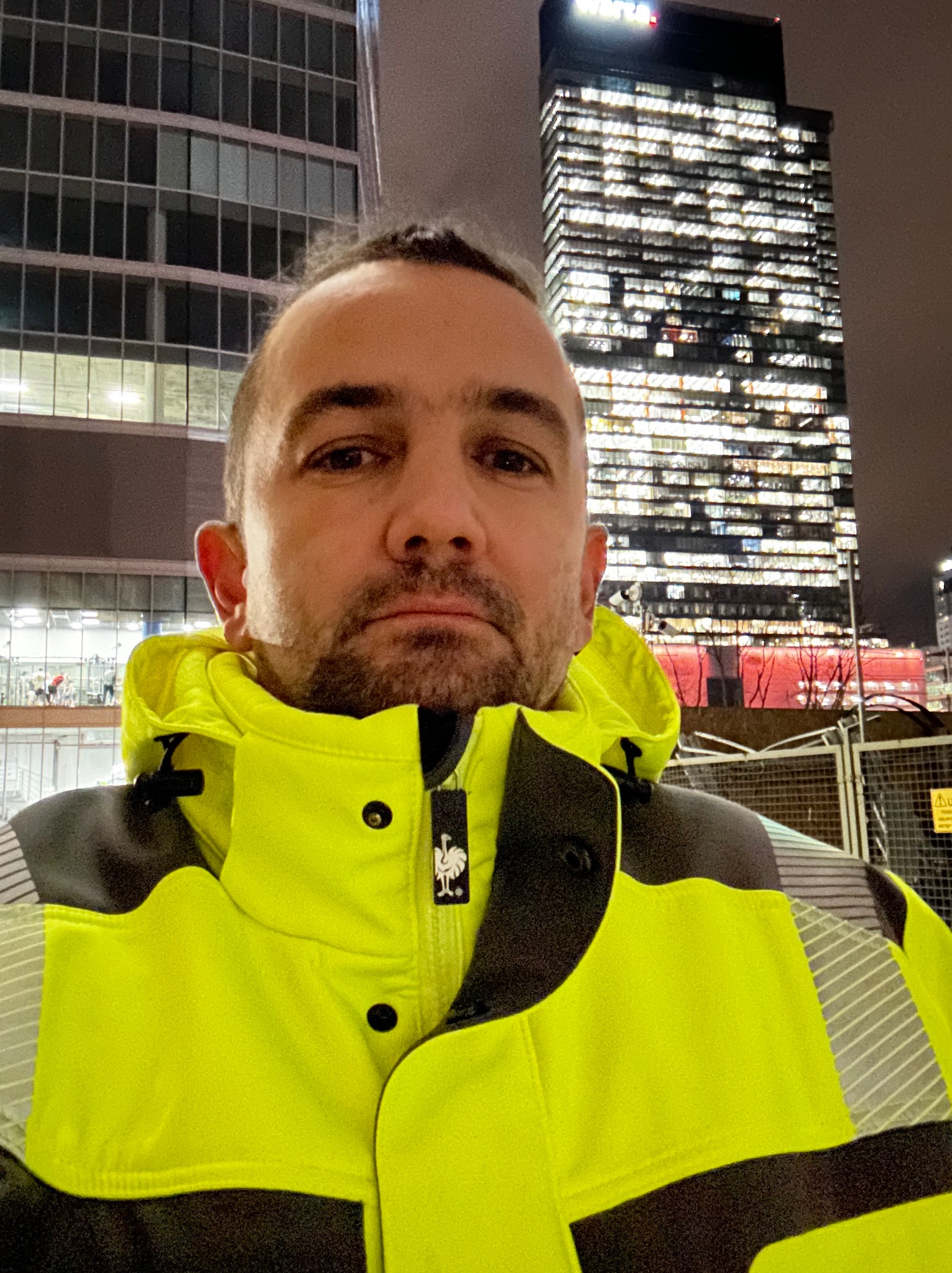 Mężczyzna w odblaskowej kurtce na tle biurowców nocą, widoczne logo Engelbert Strauss na kurtce.