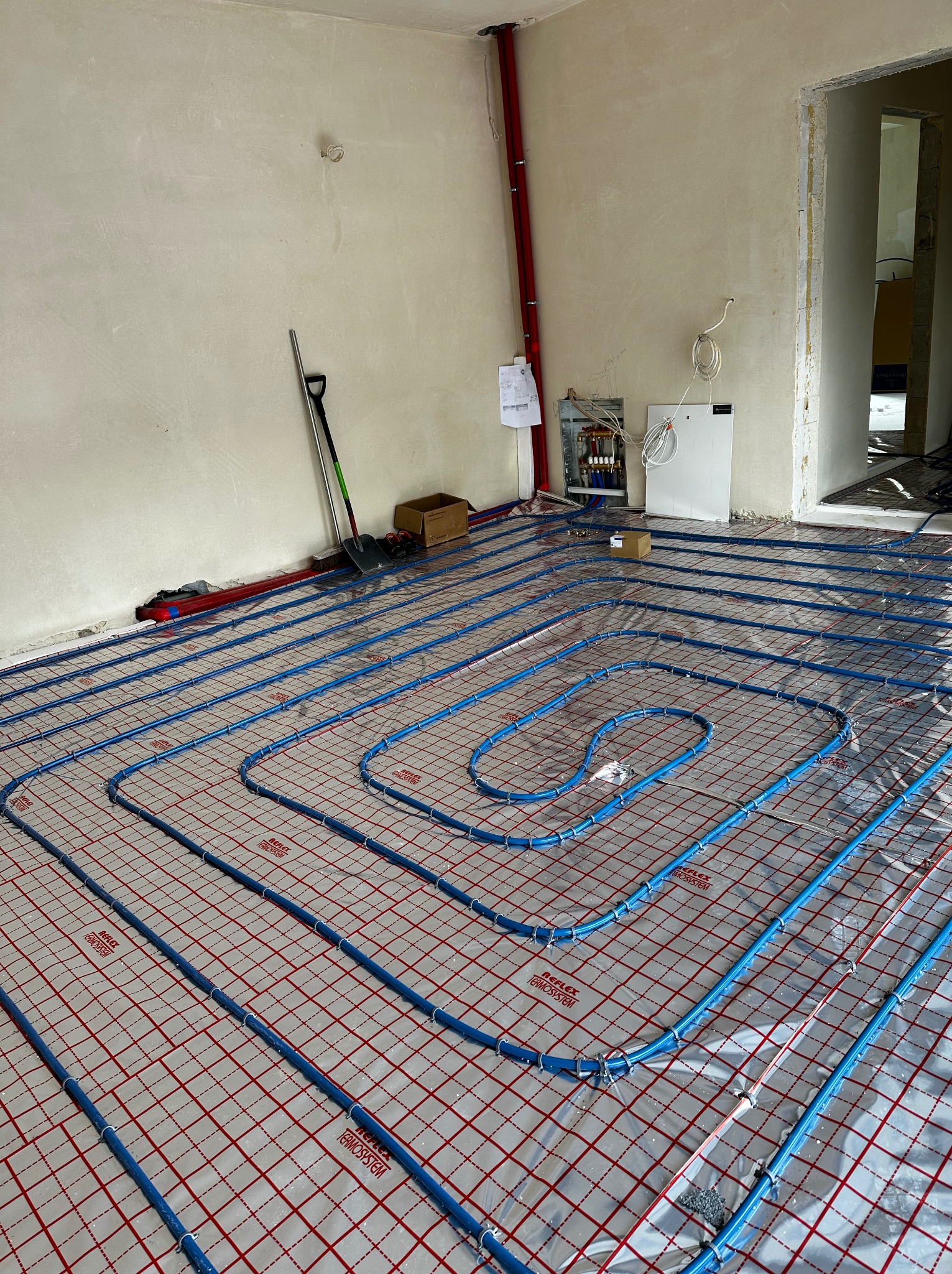 Niebieskie rury ogrzewania podłogowego rozłożone na folii z czerwoną siatką, widoczna rozdzielnia i narzędzia w surowym wnętrzu w trakcie prac instalacyjnych.