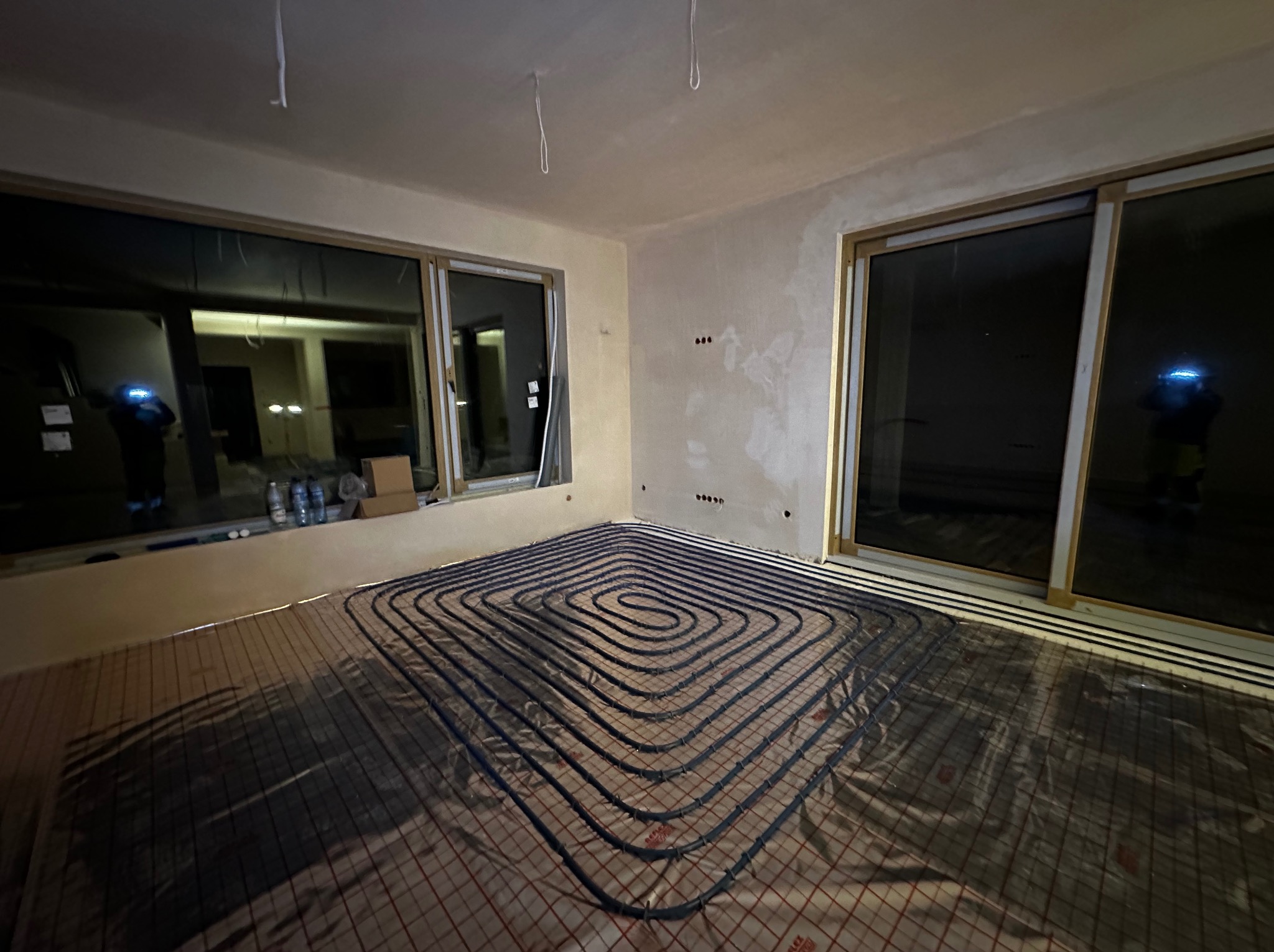 Wnętrze w trakcie prac: rozłożone rury ogrzewania podłogowego na folii z siatką, widoczne okna przesuwne i przygotowane ściany do wykończenia.