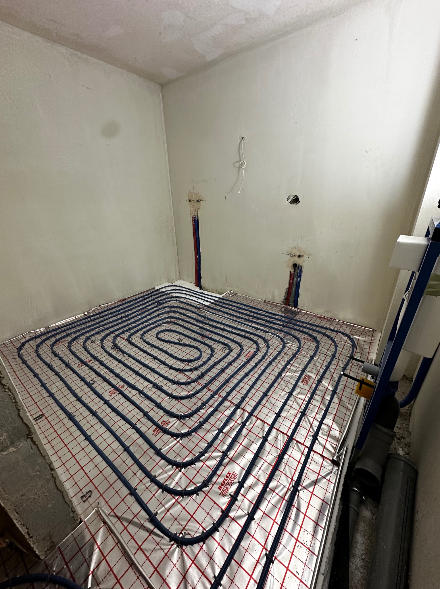 Niedokończona instalacja ogrzewania podłogowego w surowym pomieszczeniu, widoczne rury układane na folii z siatką, wystające rury instalacyjne w ścianie, brak wykończenia ścian.