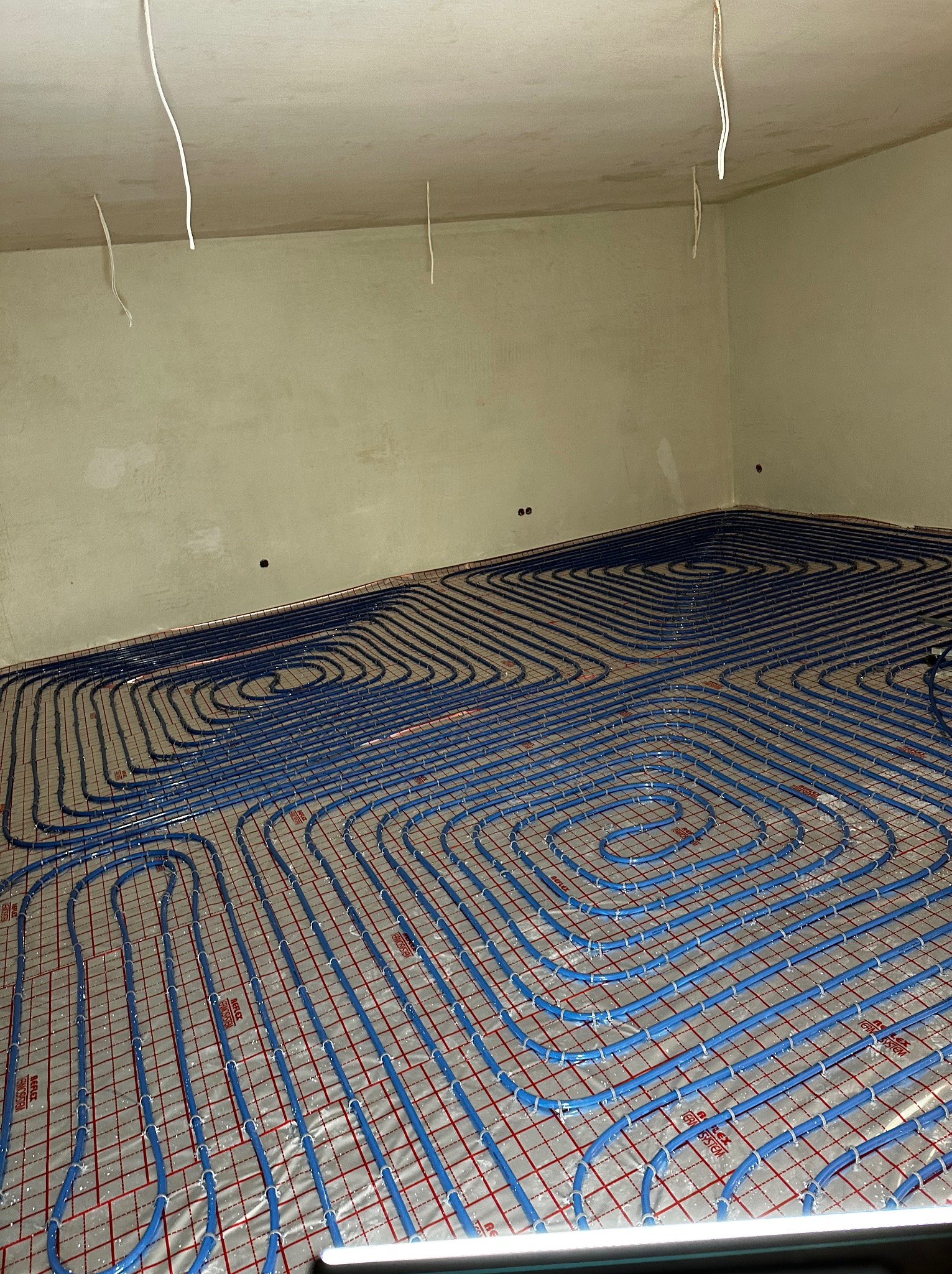 Niedokończona instalacja ogrzewania podłogowego z niebieskimi rurkami na folii z czerwoną siatką w surowym pomieszczeniu z wiszącymi kablami.