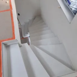 Widok z g&oacute;ry na białe, nowoczesne schody z płyt gipsowych, zabezpieczone folią malarską podczas remontu, z widocznymi śladami farby na podłodze i ścianie z cegły w tle.