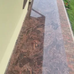 Długi, błyszczący parapet zewnętrzny z czerwono-brązowego kamienia z wyraźnym wzorem, zamontowany pod oknami budynku z żółtą elewacją, widok na zadbany ogród z zielonym trawnikiem.