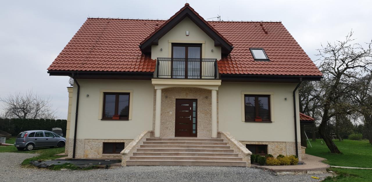 Elewacja domu jednorodzinnego z beżowymi ścianami, brązową stolarką okienną, balkonem z czarną balustradą i schodami wejściowymi wyłożonymi płytkami w odcieniach beżu.