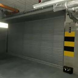 Szara, zamknięta brama rolowana w hali przemysłowej, widoczne rury wentylacyjne pod sufitem, żółto-czarne oznaczenie na ścianie oraz skrzynka z narzędziami.