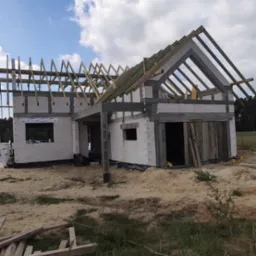 Budowa domu murowanego w trakcie realizacji: ściany z bloczków, drewniana konstrukcja dachu, teren budowy z piaskiem i zielenią.