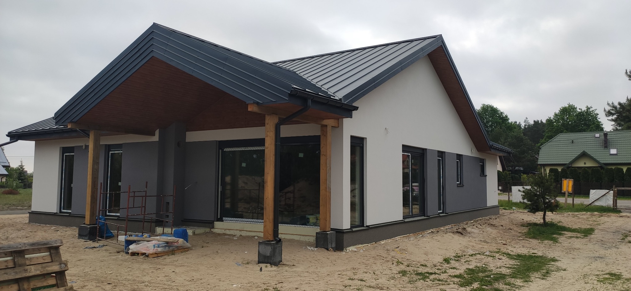 Nowoczesny dom jednorodzinny z szarą blachodachówką i drewnianym podbiciem dachu, w trakcie wykończenia elewacji. Widoczne rusztowania i materiały budowlane na piaszczystym terenie.