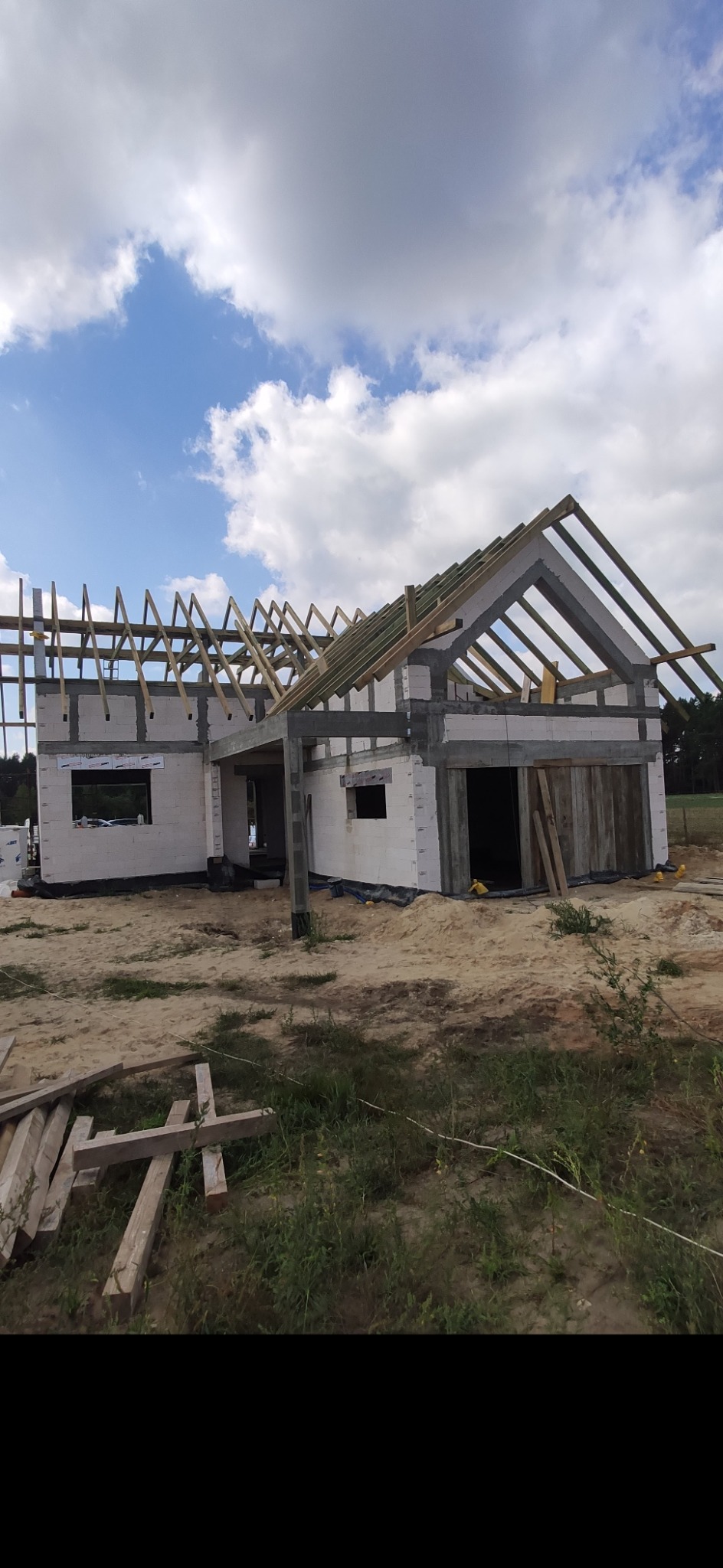 Budowa domu murowanego w trakcie realizacji: ściany z bloczków, drewniana konstrukcja dachu, teren budowy z piaskiem i zielenią.