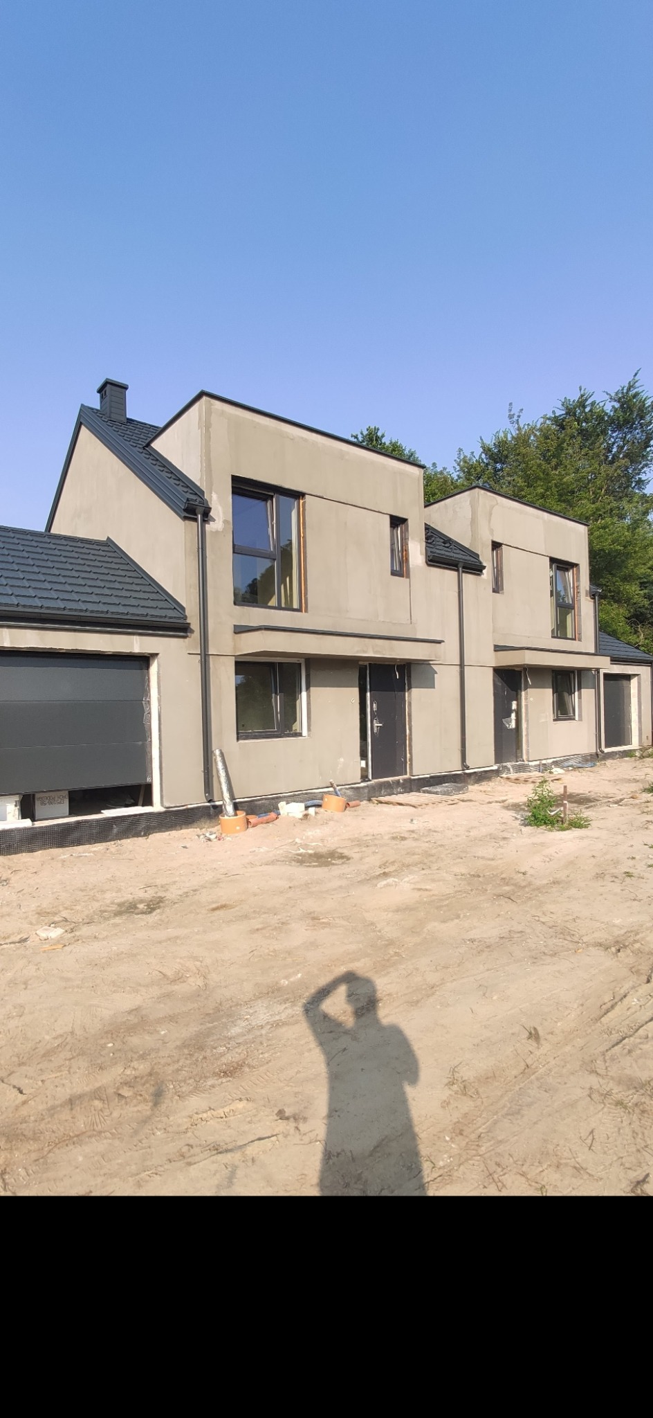 Dwa bliźniacze domy w stanie surowym zamkniętym, z szarą elewacją i ciemną dachówką, widoczne okna i drzwi, teren wokół niezagospodarowany, cień osoby robiącej zdjęcie na pierwszym planie.
