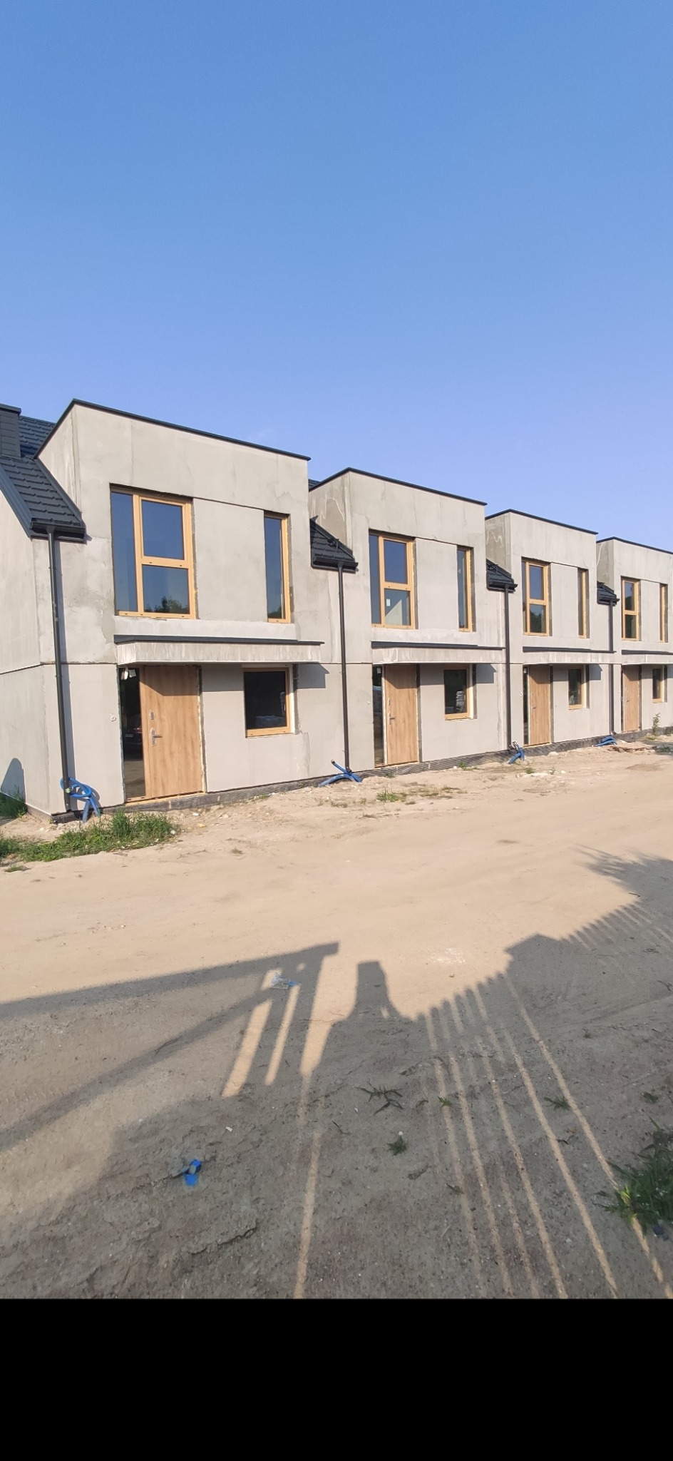 Szereg nowo wybudowanych, surowych domów w stanie deweloperskim, z drewnianymi oknami i drzwiami, na tle błękitnego nieba i piaszczystego terenu.