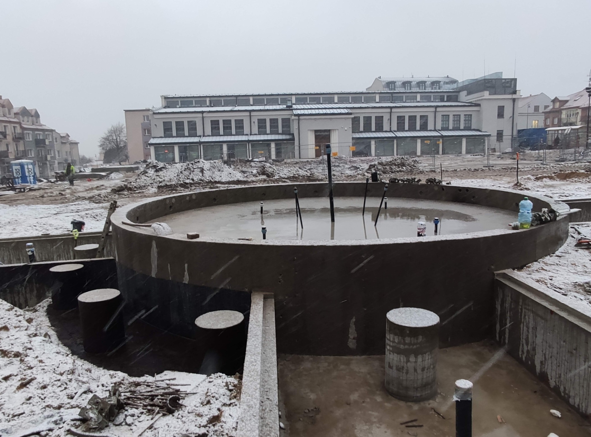 Wykop pod okrągły zbiornik zalany betonem, widoczne wystające rury i podpory, zimowa sceneria z padającym śniegiem, w tle zabudowania miejskie.
