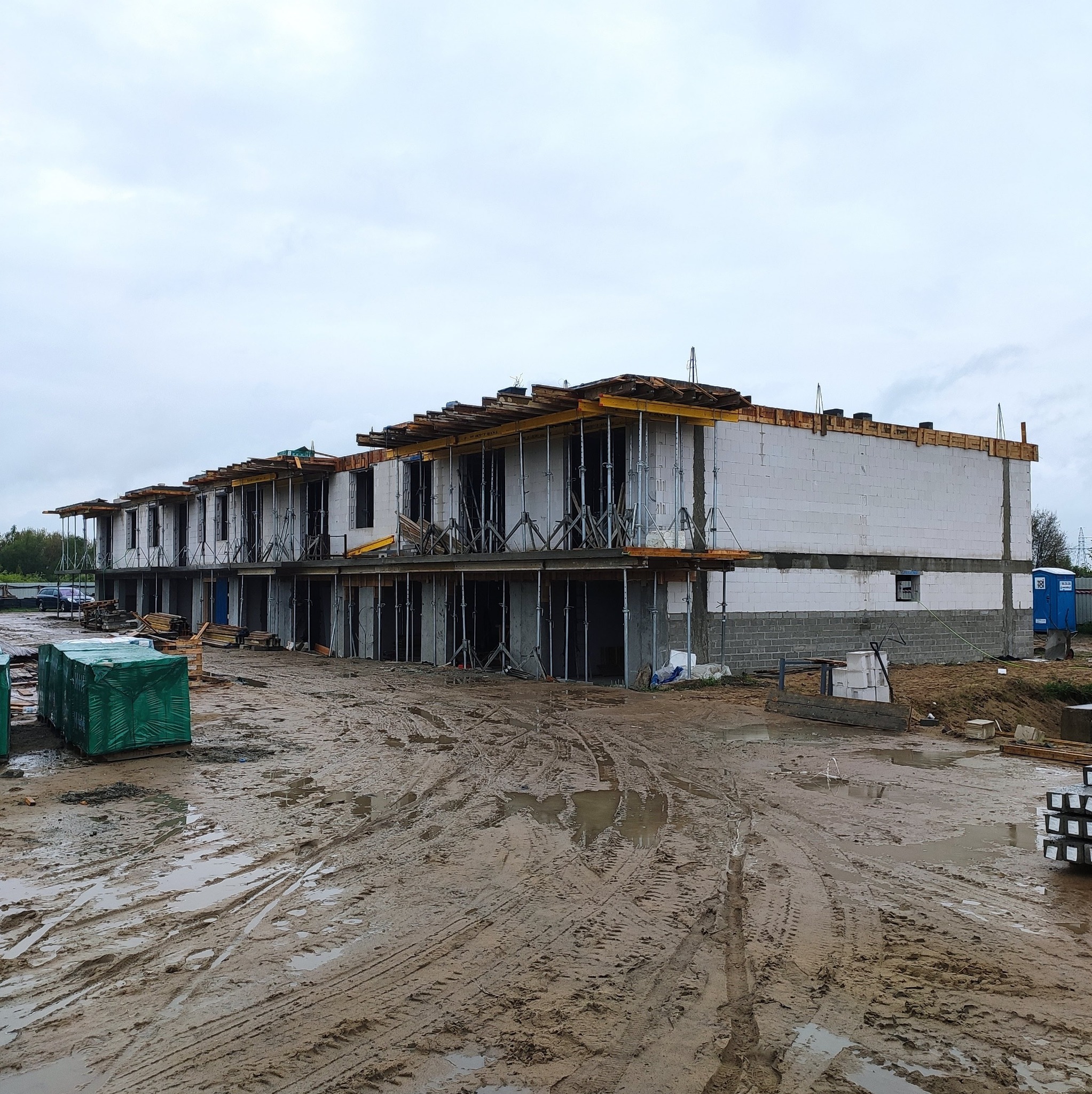 Budowa szeregu domów w stanie surowym otwartym, widoczne bloczki z betonu komórkowego, drewniane szalunki i stalowe podpory, teren budowy błotnisty po deszczu.