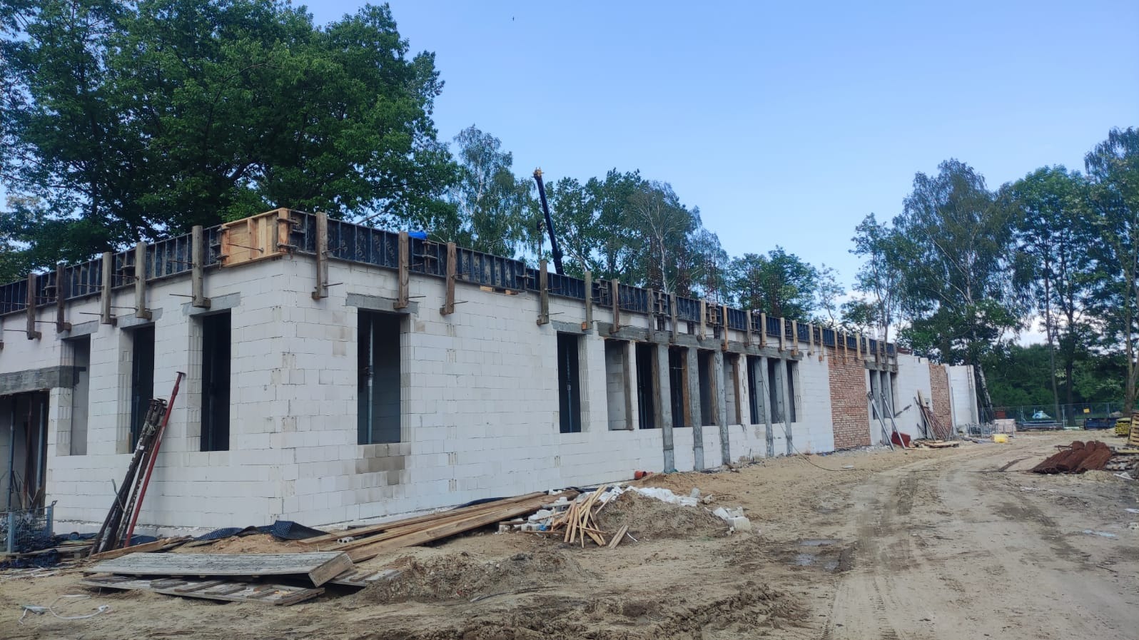 Budynek w trakcie wznoszenia z białych bloczków, z widocznymi szalunkami na górze ścian i otworami okiennymi, na tle drzew i nieba.