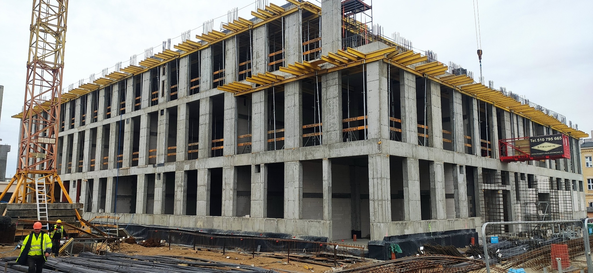 Wielopiętrowy budynek w surowym stanie, z widocznymi szalunkami stropowymi i zbrojeniami, na placu budowy z dźwigiem i pracownikami w odblaskowych kamizelkach.