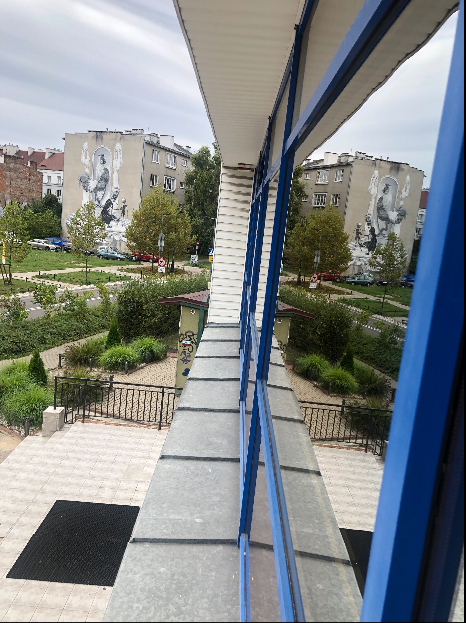 Widok z okna na budynek z muralem przedstawiającym koguta i postacie, zieleń miejską i parking, widziane przez ramę okienną z niebieskimi elementami.