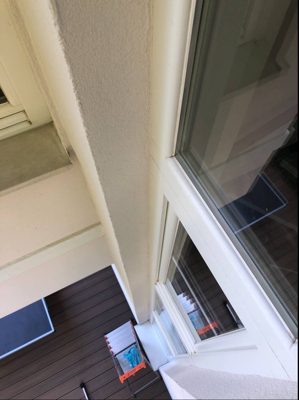 Widok z góry na okno z białą ramą, część elewacji z fakturą baranka, aluminiową drabinę opartą o ścianę i fragment tarasu z desek kompozytowych