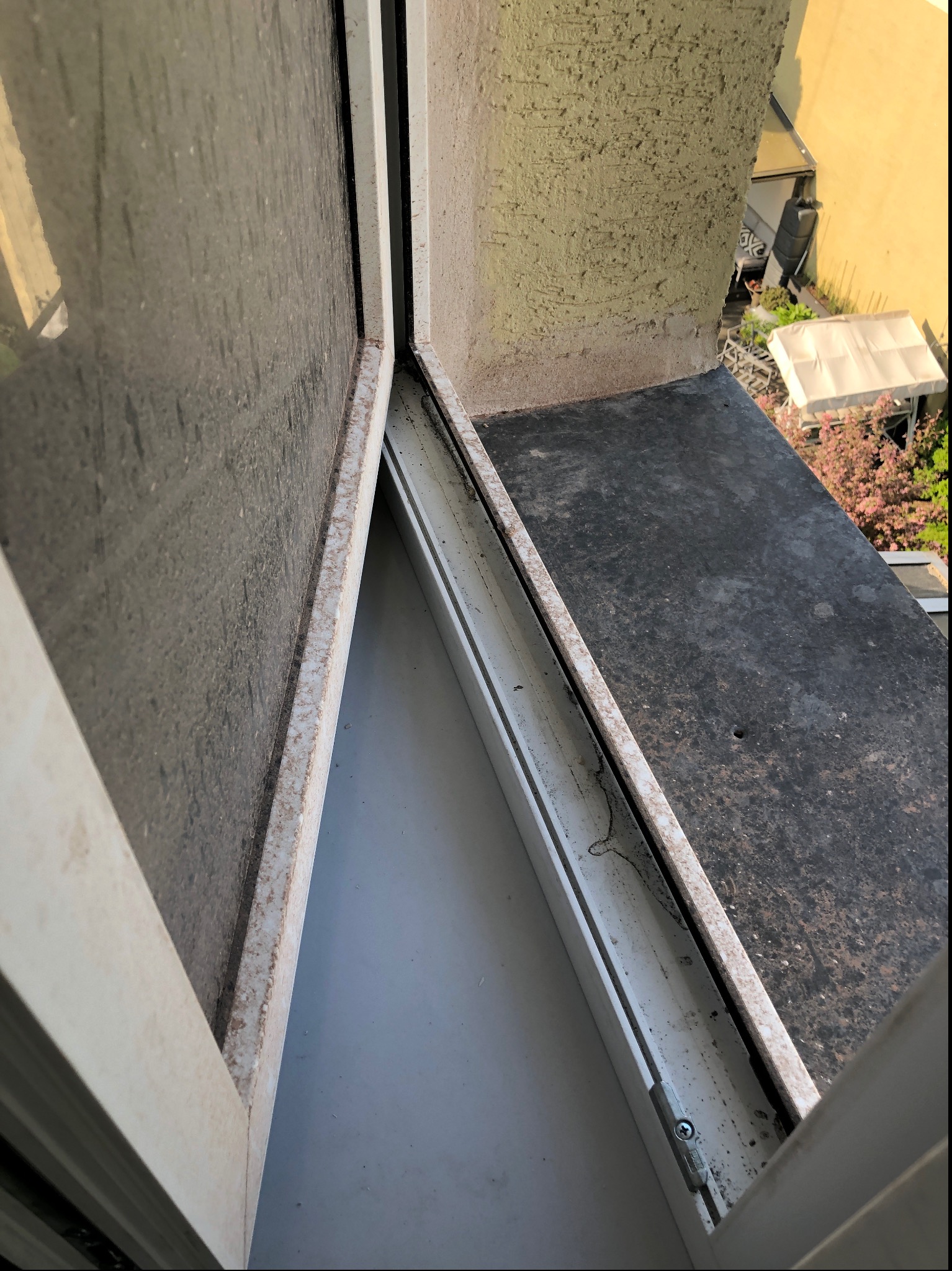 Silnie zabrudzone okno balkonowe, widok z góry, z widocznymi śladami kurzu i zabrudzeń na szybie, ramie okiennej oraz wnęce okiennej. Na zewnątrz widoczny fragment balkonu i zieleni.