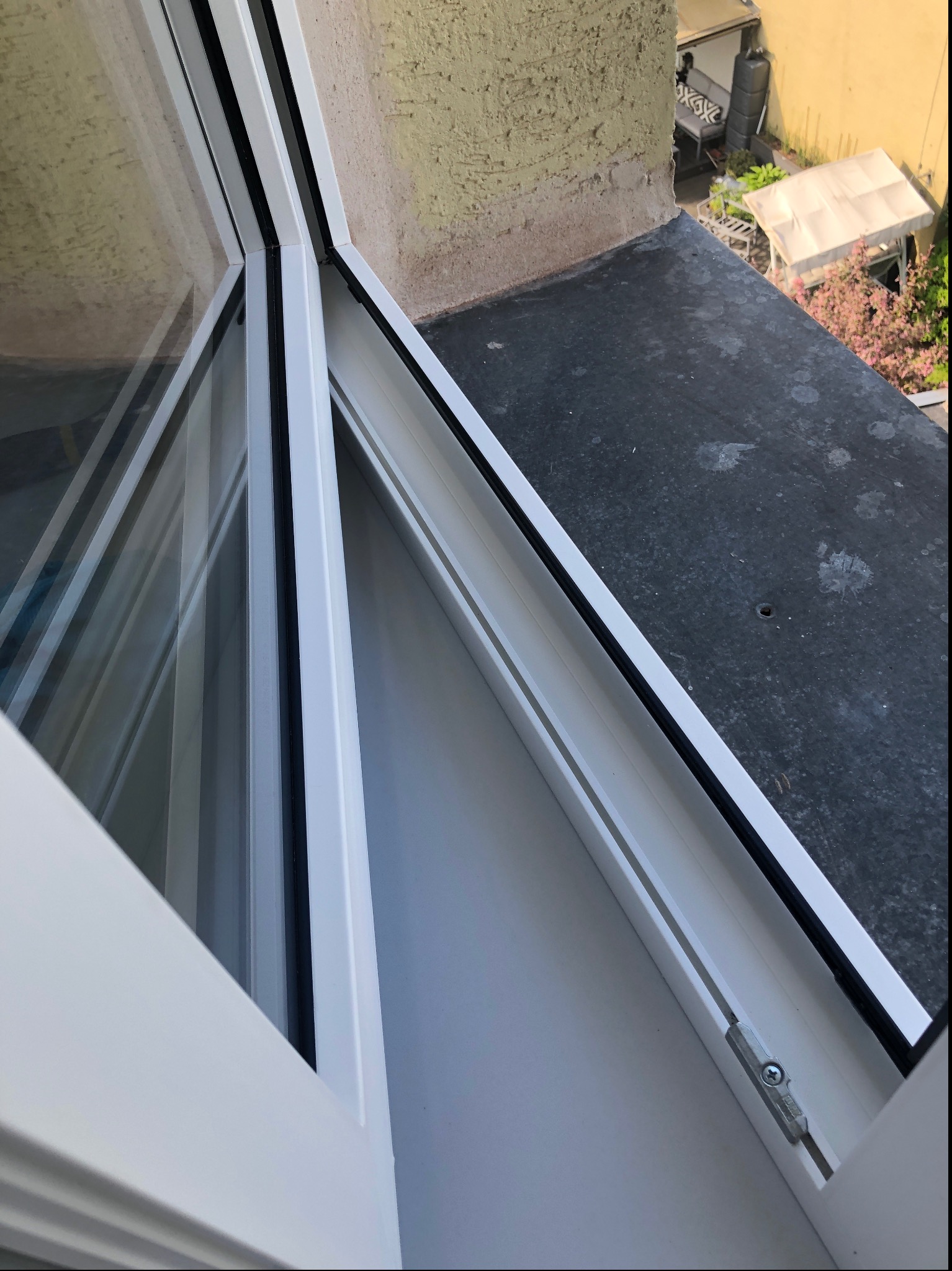Nowe, białe okno PCV z widocznym mechanizmem blokującym, uchwycone z bliska pod kątem, z widokiem na czarny parapet zewnętrzny i fragment ogrodu.
