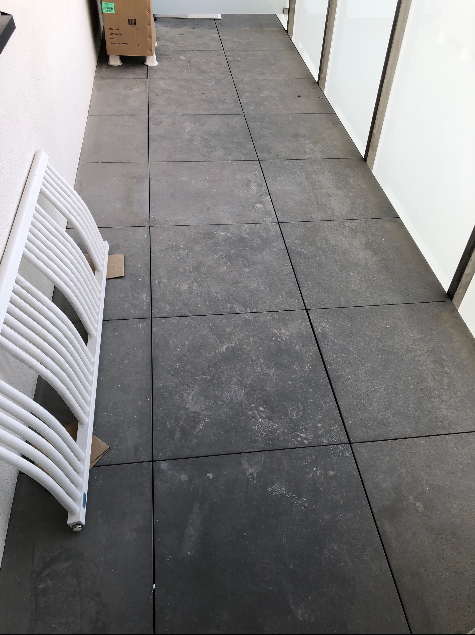 Szare płytki podłogowe po remoncie z widocznymi śladami stóp i kartonem oraz białym grzejnikiem żeberkowym opartym o ścianę.