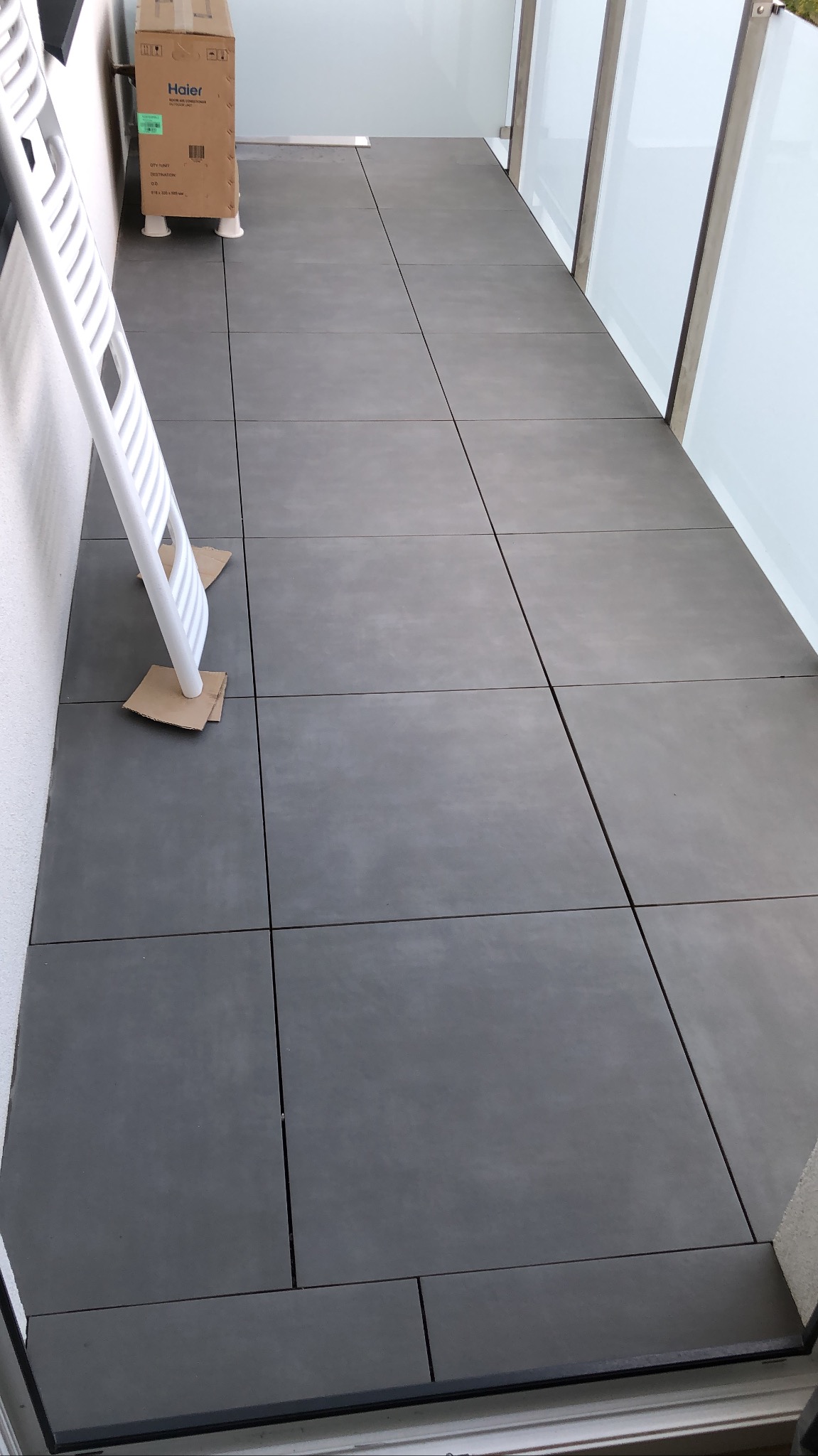 Szare płytki na balkonie po remoncie, zabezpieczone kartonem pod drabiną, z widocznym kartonem po urządzeniu AGD.