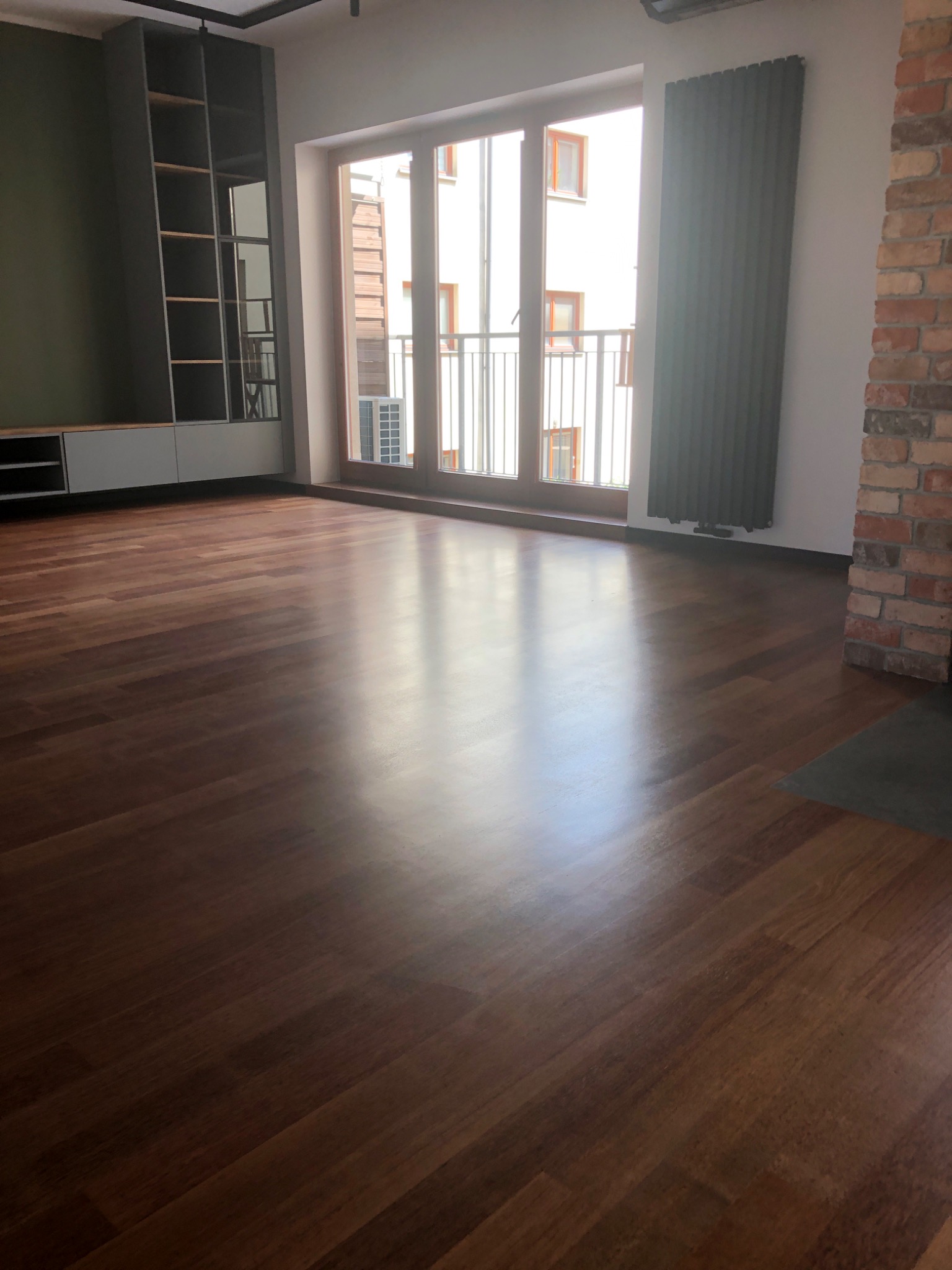 Wnętrze pokoju po remoncie z parkietem w odcieniach brązu, widoczne okno balkonowe z widokiem na budynek, szara zabudowa meblowa z półkami oraz fragment ściany z cegły.