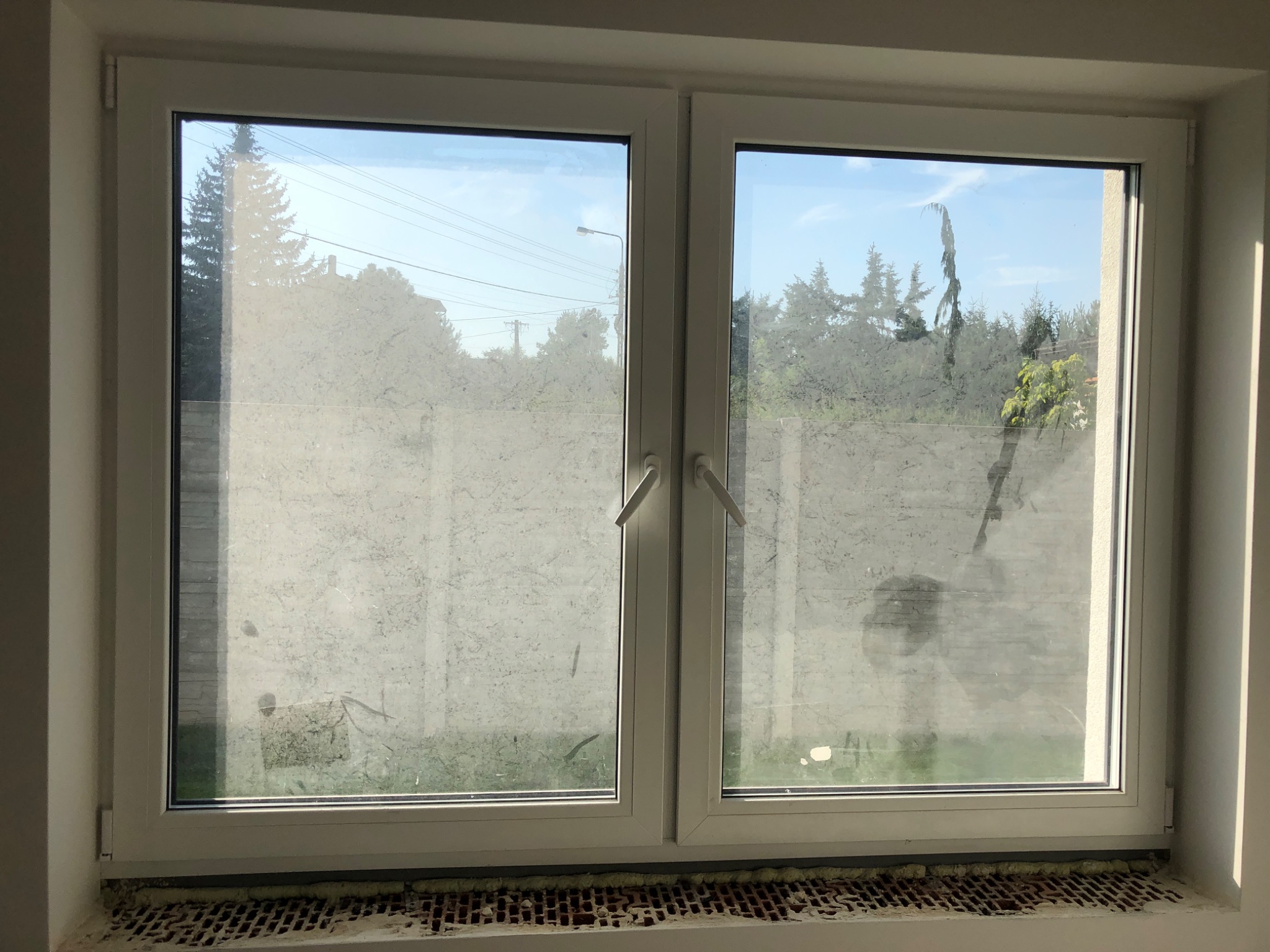 Białe, dwuskrzydłowe okno PCV, z widocznymi zabrudzeniami po remoncie na szybach, przez które widać drzewa i niebieskie niebo. W dolnej części okna widoczny mur z cegieł wypełniony pianką montażową.