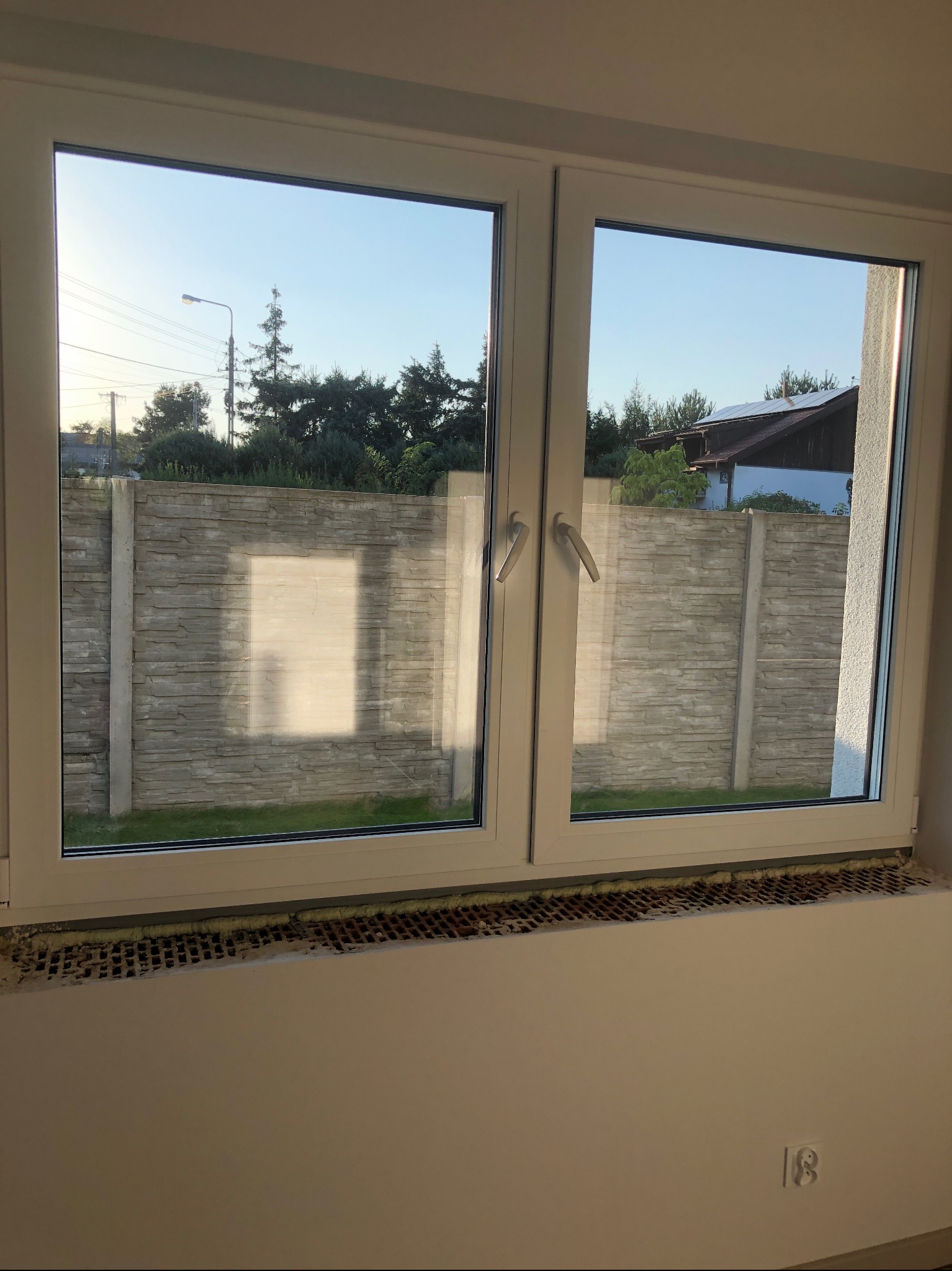 Widok przez nowe, dwuskrzydłowe okno PCV na betonowy płot i zieleń pod błękitnym niebem; widoczne ślady pianki montażowej i pozostałości po pracach budowlanych na parapecie.