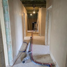 Lukas-Tynk Tynki Maszynowe Łukasz Tchoryk - Wnętrze korytarza w trakcie prac wykończeniowych: ściany pokryte świeżym, jasnym tynkiem, widoczne instalacje sanitarne na podłodze oraz surowy strop z widocznymi przewodami oświetleniowymi.