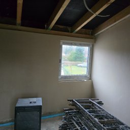 Lukas-Tynk Tynki Maszynowe Łukasz Tchoryk - Wnętrze w trakcie prac wykończeniowych: ściany pokryte świeżym tynkiem, widoczne rusztowanie, grzejnik elektryczny i okno z widokiem na zieleń.