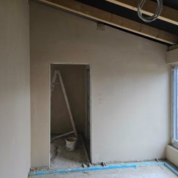 Lukas-Tynk Tynki Maszynowe Łukasz Tchoryk - Wnętrze w trakcie prac wykończeniowych: świeżo otynkowane ściany w jasnym odcieniu, widoczne drewniane elementy konstrukcyjne sufitu oraz okno z widokiem na zewnątrz, drabina oparta o ścianę...