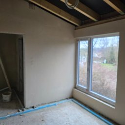 Lukas-Tynk Tynki Maszynowe Łukasz Tchoryk - Wnętrze pokoju po nałożeniu świeżego tynku, widoczne drewniane elementy stropu, okno z widokiem na zewnątrz oraz niebieskie rurki zabezpieczające podłogę.