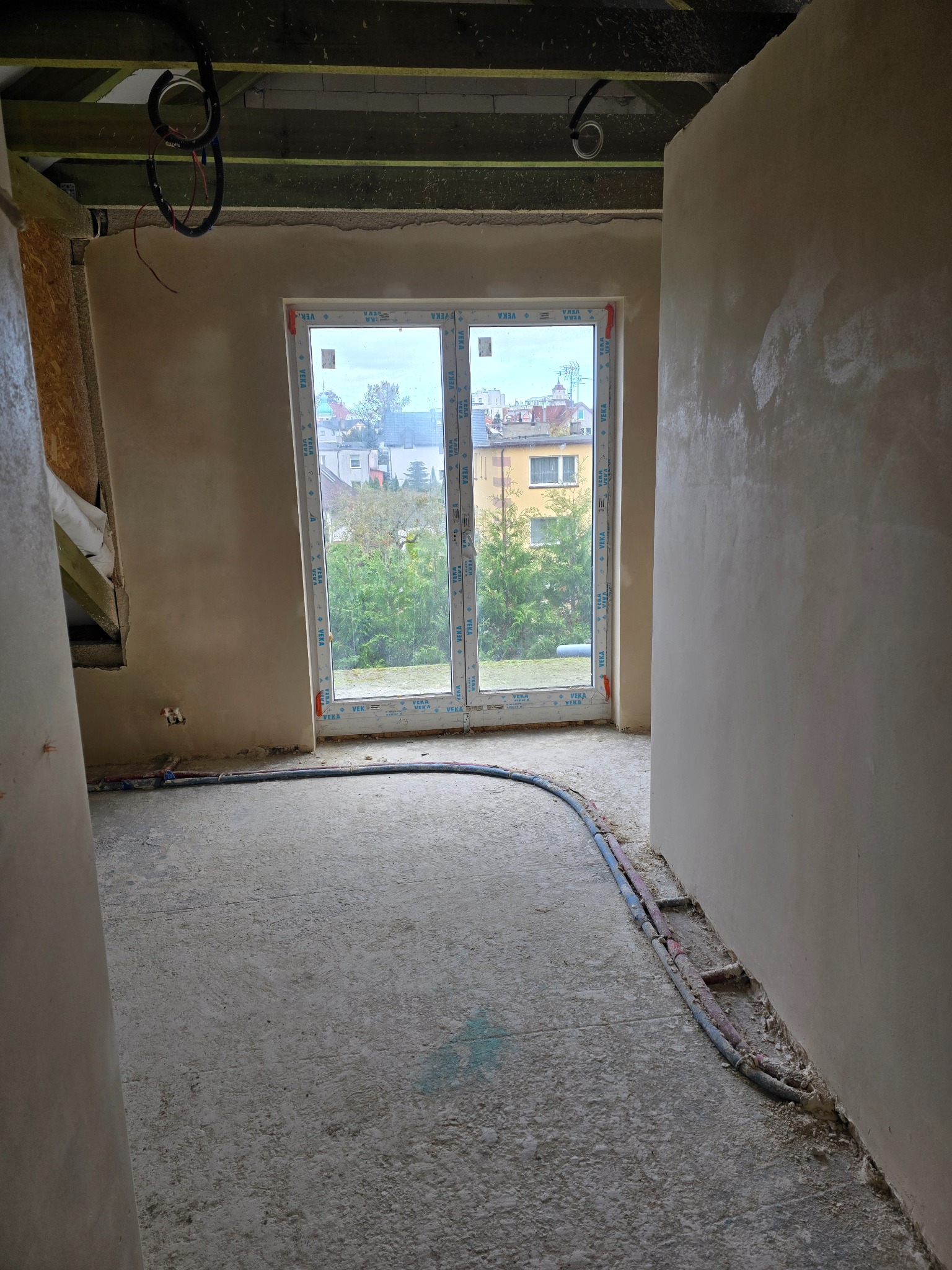 Surowe wnętrze w trakcie prac wykończeniowych: ściany pokryte świeżym tynkiem, widoczne przewody instalacji grzewczej w podłodze, okno z widokiem na zabudowę miejską i zieleń. Na suficie widoczne...