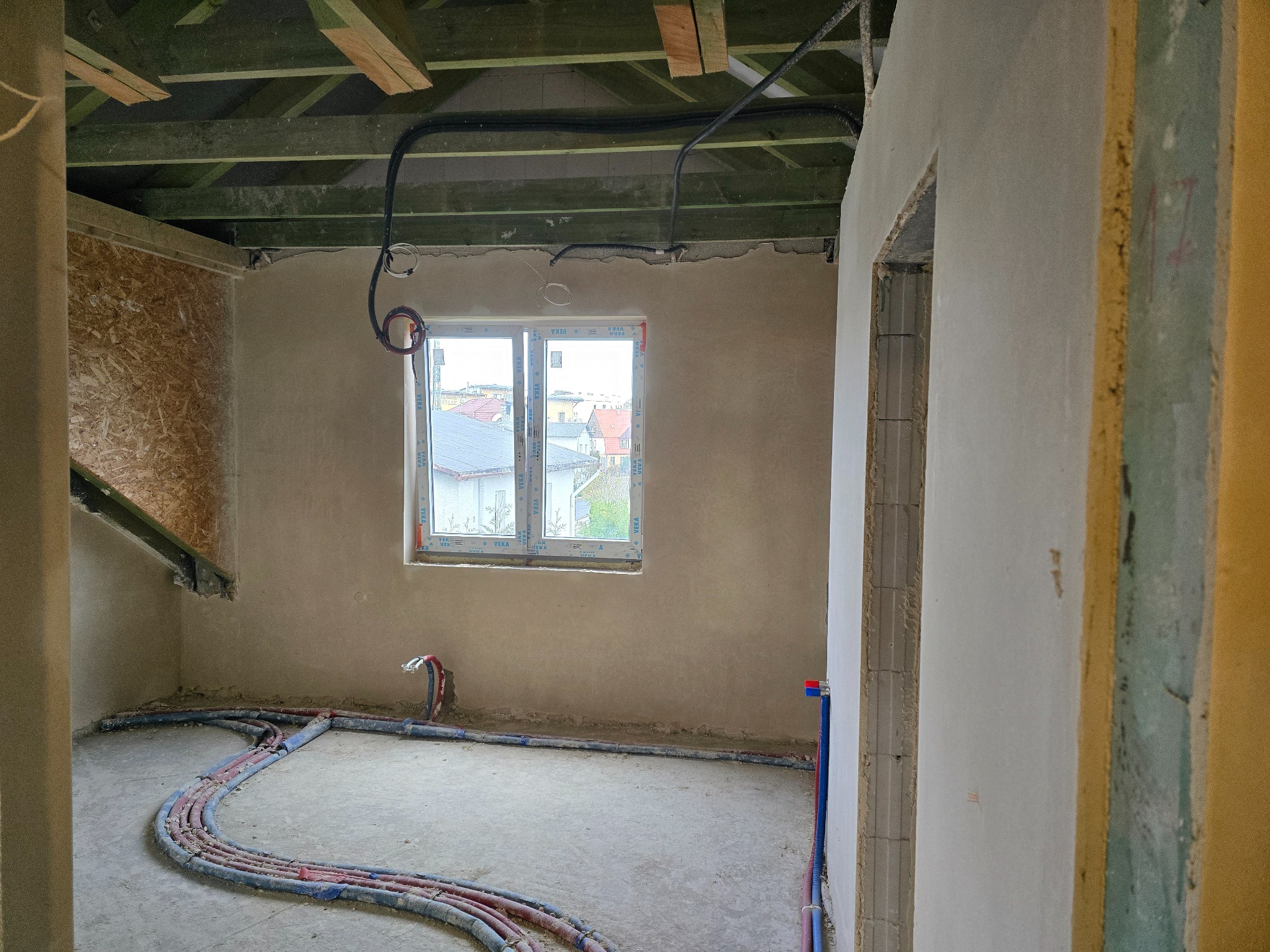 Surowy stan wnętrza: ściany po nałożeniu wstępnej warstwy tynku, widoczne rury instalacji podłogowej, sufit z drewnianą konstrukcją i przewodami elektrycznymi.