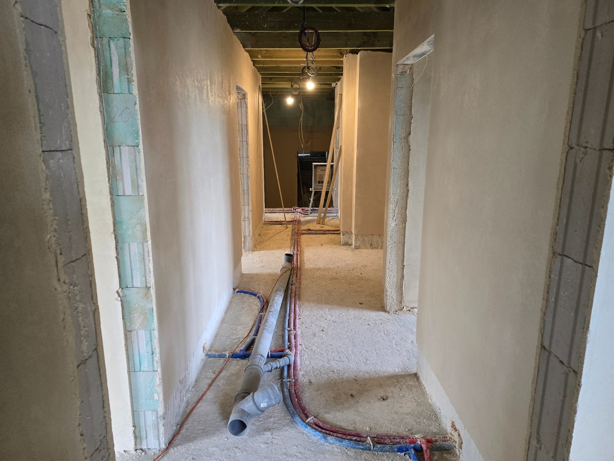 Wnętrze korytarza w trakcie prac wykończeniowych: ściany pokryte świeżym, jasnym tynkiem, widoczne instalacje sanitarne na podłodze oraz surowy strop z widocznymi przewodami oświetleniowymi.