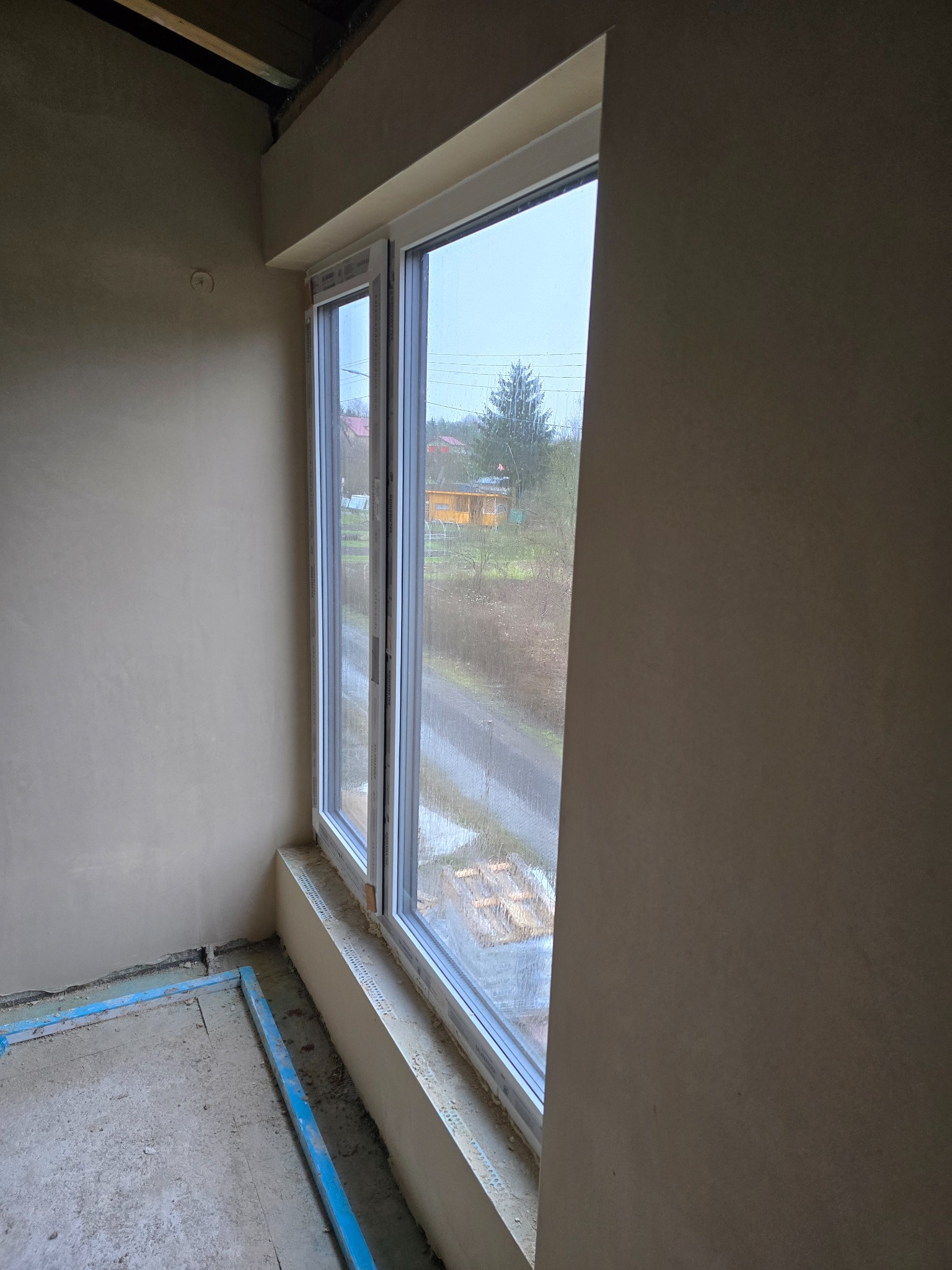 Wnętrze pokoju w trakcie prac wykończeniowych, ściany pokryte świeżym, jasnym tynkiem, widoczne nowe okno PCV z widokiem na zewnątrz w pochmurny dzień i fragment ramy pod wylewkę podłogi.