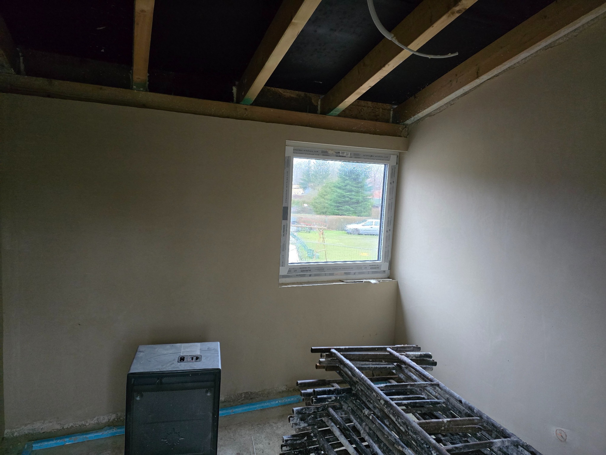 Wnętrze w trakcie prac wykończeniowych: ściany pokryte świeżym tynkiem, widoczne rusztowanie, grzejnik elektryczny i okno z widokiem na zieleń.