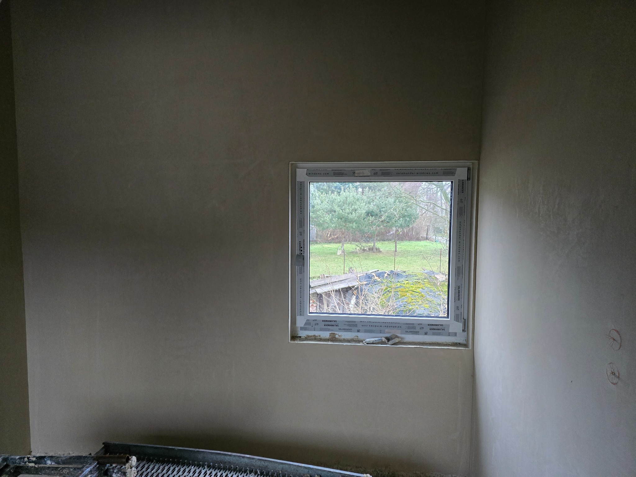 Wnętrze z oknem po nałożeniu świeżego tynku, widoczna szpachelka i drabina malarska w dolnej części kadru.