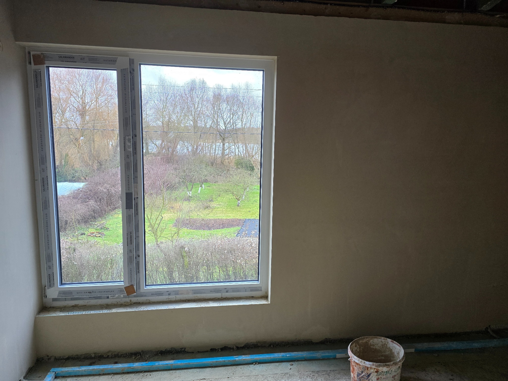 Świeżo otynkowana ściana wewnętrzna z widocznym oknem Salamander i elementami rusztowania podłogowego, widok na ogród z drzewami bez liści.