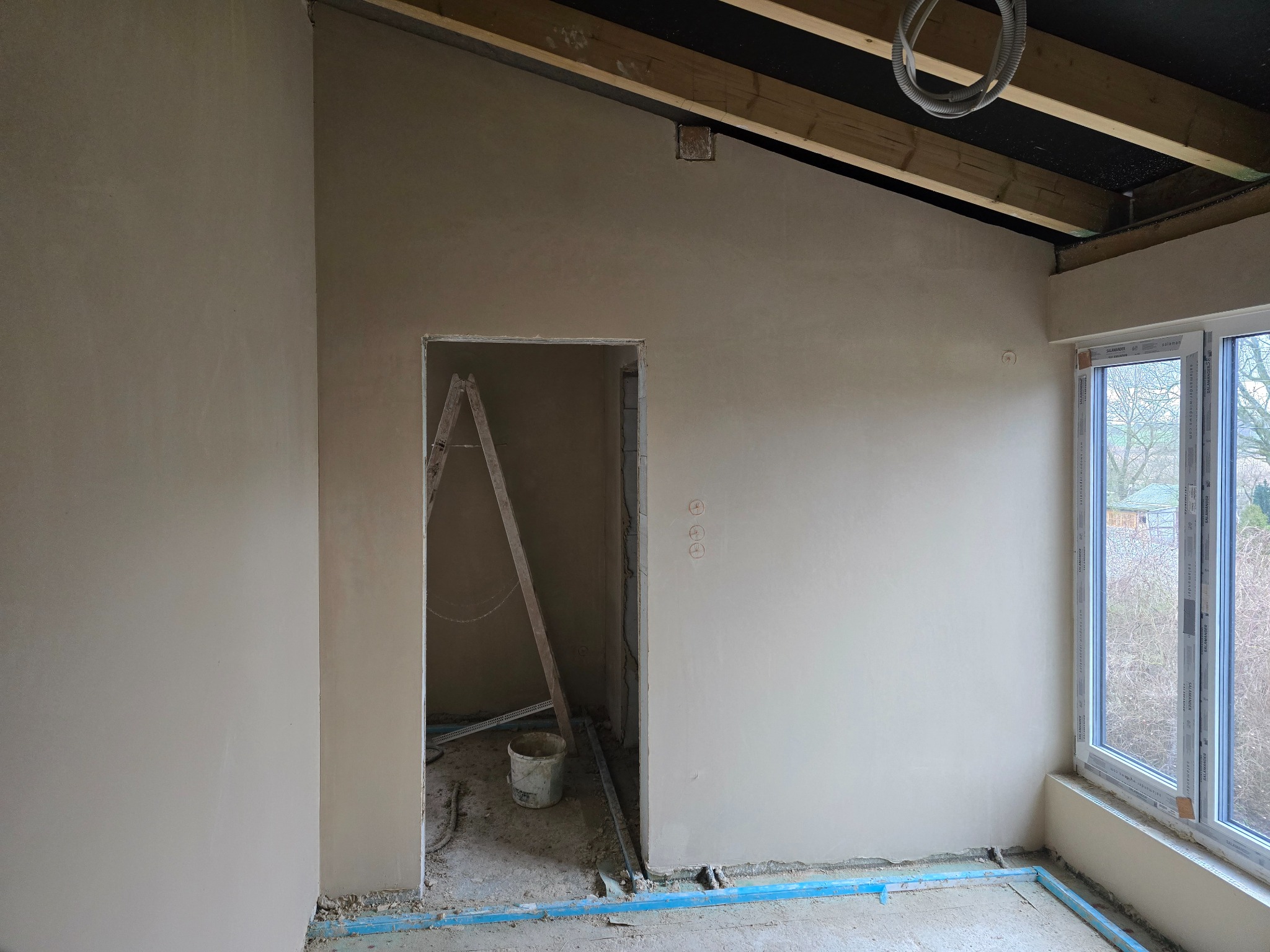 Wnętrze w trakcie prac wykończeniowych: świeżo otynkowane ściany w jasnym odcieniu, widoczne drewniane elementy konstrukcyjne sufitu oraz okno z widokiem na zewnątrz, drabina oparta o ścianę...