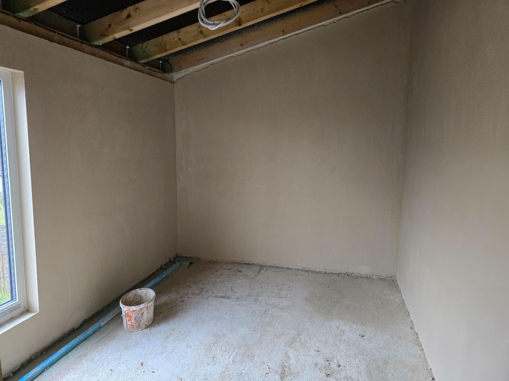 Pokój po nałożeniu gładzi gipsowej na ściany, widoczny fragment okna, drewniana konstrukcja sufitu, wiadro i niebieska rurka przy ścianie.