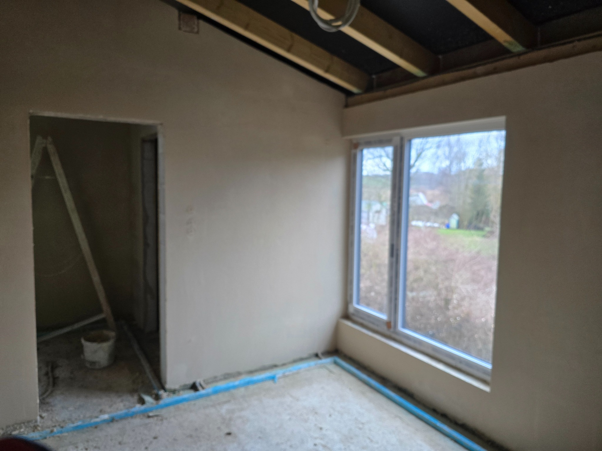 Wnętrze pokoju po nałożeniu świeżego tynku, widoczne drewniane elementy stropu, okno z widokiem na zewnątrz oraz niebieskie rurki zabezpieczające podłogę.