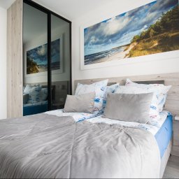 PRZEMYSŁAW JASZCZYK - Jasna sypialnia z łóżkiem w szaro-niebieskiej tonacji, nad nim obraz z motywem morza. W tle szafa z lustrzanymi drzwiami.